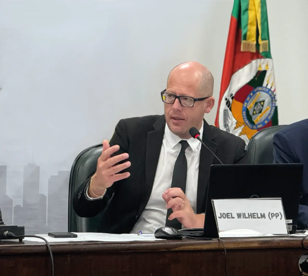 Joel Wilhelm falando ao microfone na mesa de uma comissão, com a bandeira do Rio Grande do Sul ao fundo