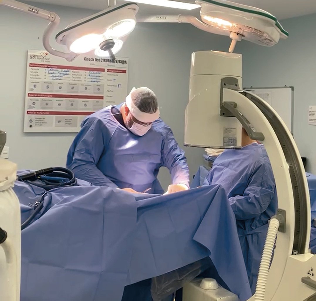 Dois profissionais de avental azul em cirurgia, com focos cirúrgicos acesos e aparelho de arco em C ao lado da mesa.