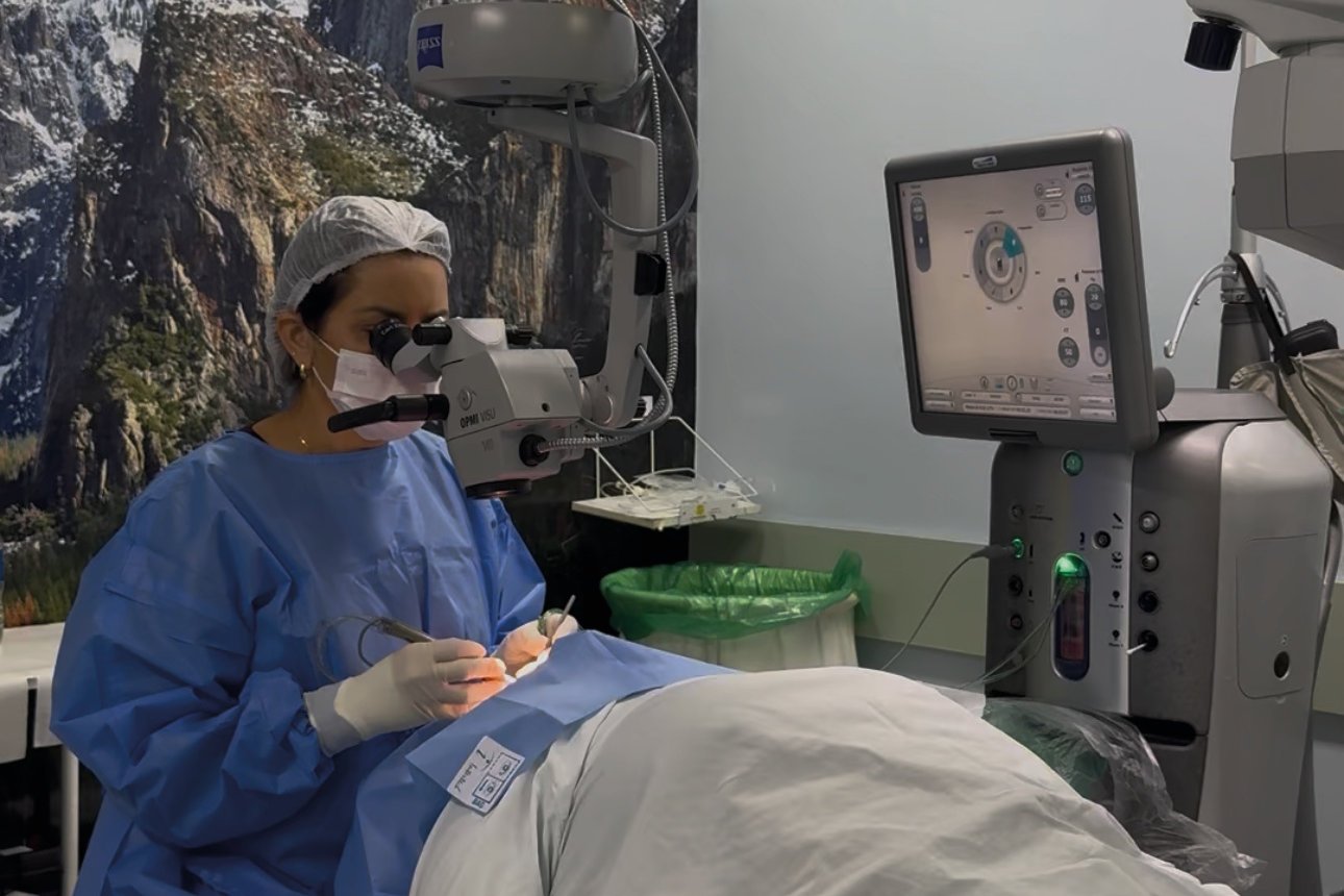 Cirurgiã de avental azul operando com microscópio cirúrgico em uma sala de cirurgia oftalmológica, com monitor do equipamento ao lado.