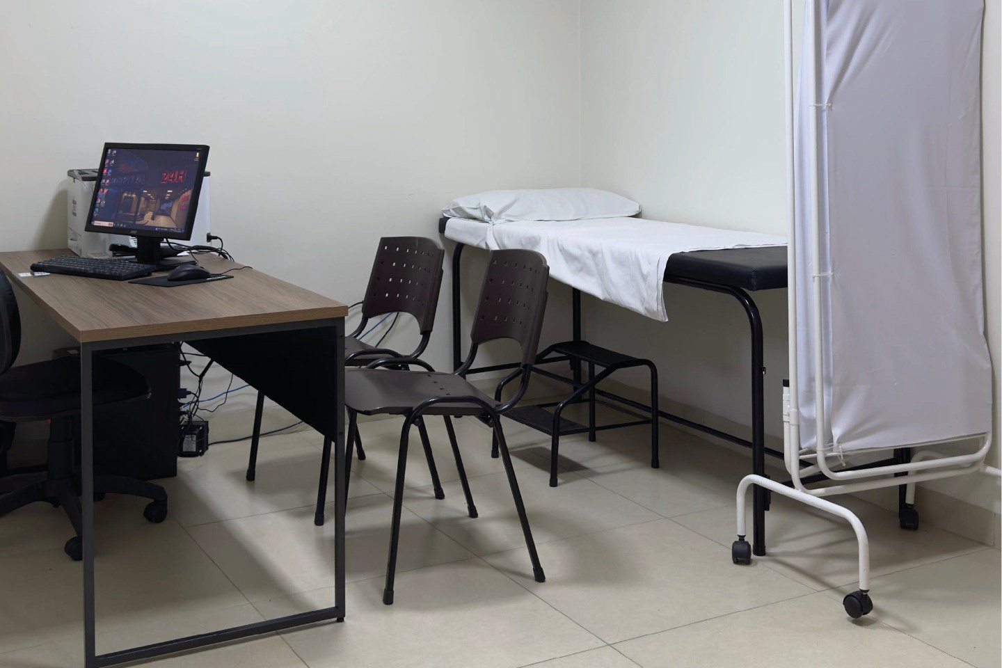 Consultório com mesa de trabalho, computador, cadeiras, maca de exame e biombo com rodas.