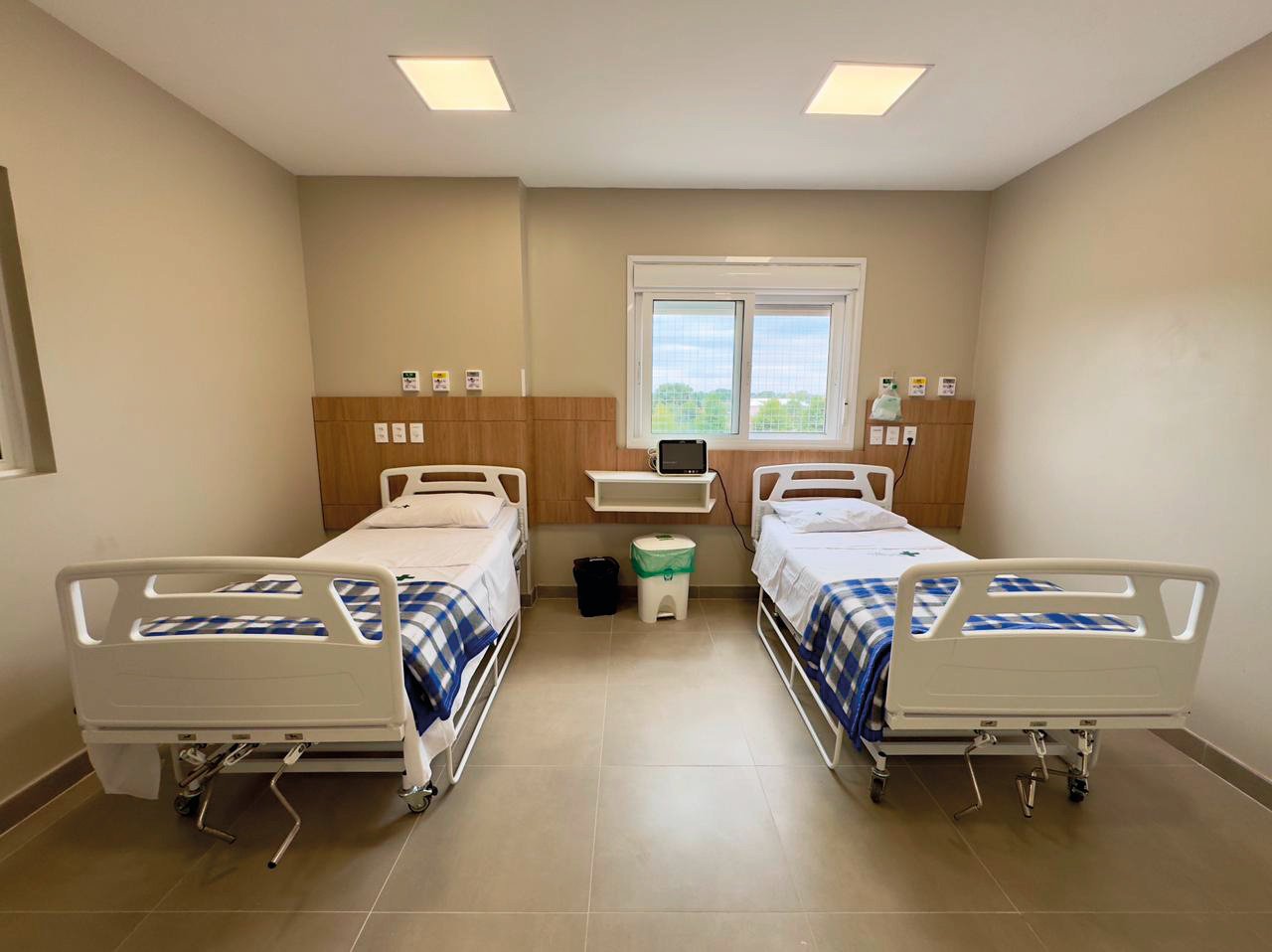 Quarto de hospital com dois leitos elétricos, janela ao fundo e painel de madeira na parede.